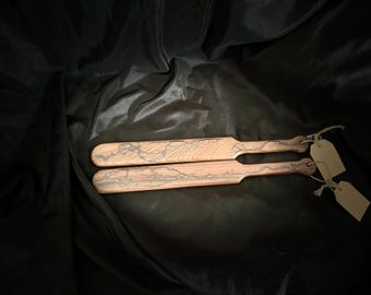 May include: Two wooden paddles with a light brown finish and a dark gray lightning bolt design. The paddles have a handle and a tag attached with twine. The background is a dark, textured fabric.