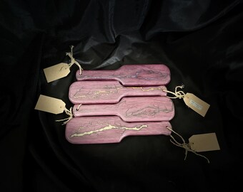 May include: A collection of four paddle-shaped wooden serving boards. Each board is a vibrant pink with a unique, lightning-like design in gold and gray. Blank tags are attached with twine, suggesting they are for sale.