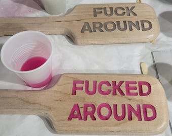 May include: Two wooden paddles with the words "FUCK AROUND" and "FUCKED AROUND" carved into them. The letters are filled with pink resin. A plastic cup with pink liquid is in the foreground.