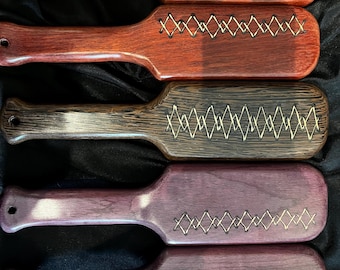 May include: Five wooden paddles in varying shades of brown and purple. Each paddle has a handle with a hole and a rectangular head. The heads are decorated with a zig-zag pattern made of a lighter-colored material.