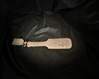 May include: A wooden paddle with a light brown finish and a leopard print design. The paddle has a handle and a rectangular head. A tag is attached to the handle with twine. The paddle is on a black background.