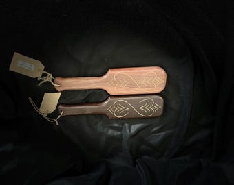May include: Two wooden paddles with decorative etched designs. One paddle is light brown, the other is dark brown. Both have a handle and a rectangular head with an etched design. The paddles are displayed on a black surface.