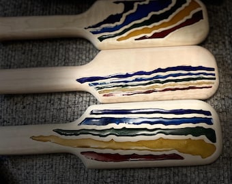May include: Three wooden paddles with colorful, layered designs. Each paddle features a light wood finish with bands of blue, green, yellow, and red. The designs resemble abstract landscapes or waves, adding a decorative touch to the paddles.