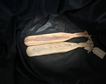 May include: Two wooden paddles with decorative burned designs. One paddle is light brown with red accents, the other is a lighter wood tone. Both have a natural wood grain and are tied together with twine and a tag.