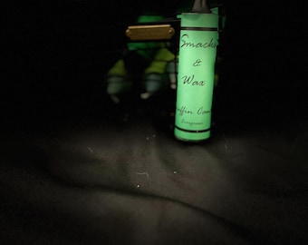 May include: A green glass jar with a black label that reads "Snacks & Wax" and "Muffin Candle" in white text. The jar is glowing in the dark.