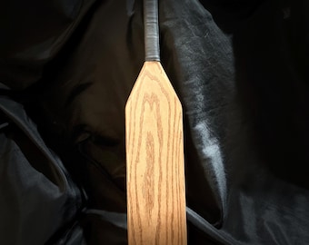 May include: A wooden paddle with a black leather handle and a circular wooden knob at the top. The paddle has a natural wood finish and a grain pattern.