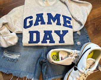 Può includere: Una felpa grigio chiaro con la scritta "GAME DAY" in lettere blu glitterate è abbinata a pantaloncini di jeans e scarpe da ginnastica bianche con dettagli neri. L'outfit è disposto su una superficie di legno.