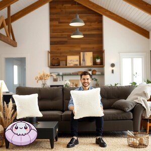 Op de afbeelding: Een man zit op een bruine bank met twee witte kussens. Hij houdt een wit kussen voor zich. De bank staat in een woonkamer met een houten plafond en een houten muur achter de bank.