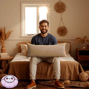 Op de afbeelding: Een man zit op een bed en houdt een beige kussen voor zich. Het bed heeft een bruin dekbed en witte kussens. De kamer heeft een raam met zonlicht dat erdoorheen schijnt en een houten bijzettafel met een lamp.