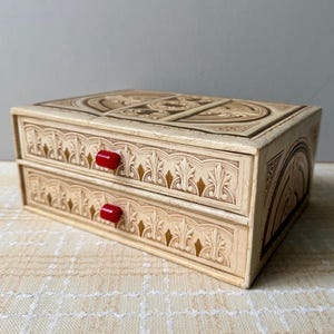 May include: A rectangular cream-colored storage box featuring two drawers with bright red cylindrical knobs. The exterior surfaces are decorated with intricate embossed patterns, including arched motifs and gold-toned accents.