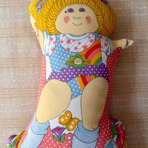 May include: A fabric doll with yellow yarn-style hair, purple eyes, and pink cheeks. The doll wears a blue and white star-patterned top, a purple polka-dot sash, and blue leg warmers. The background fabric features a rainbow and floral print.