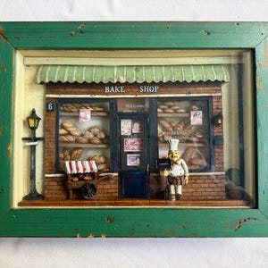 Puede incluir: Diorama enmarcado de una panadería con un marco de madera verde desgastada. La tienda tiene un toldo verde, fachada de ladrillo y una figura de panadero en miniatura. La tienda exhibe varios productos horneados y tiene las palabras "BAKE SHOP".