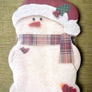 May include: A snowman-shaped decorative item featuring a red hat with a white textured brim, a plaid scarf, a small heart patch, and a red bird at the base. The snowman has black bead eyes and an orange nose. The text CURRENT, INC. is printed at the bottom.