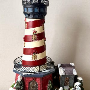 May include: A decorative lighthouse figurine featuring red and white horizontal stripes, a black lantern room, and a red brick base. The structure is adorned with small holiday wreaths and patches of white snow, set upon a base of sculpted gray waves.