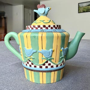May include: A ceramic teapot featuring vertical yellow and green stripes, decorated with raised light blue bird figures. The lid is topped with a blue bird and a red heart, and the rim displays a red and white checkerboard pattern with numbers.
