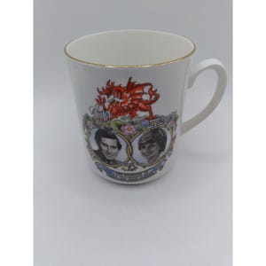 May include: A white ceramic mug featuring a gold rim and a commemorative graphic celebrating the 1981 marriage of the Prince of Wales and Lady Diana Spencer. The design includes a red dragon, floral accents, and portraits of the couple.