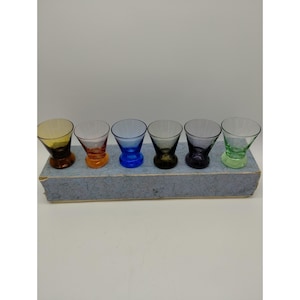 Pode incluir: Um conjunto de seis copos pequenos em várias cores, incluindo âmbar, laranja, azul, castanho escuro, roxo e verde claro. Cada copo apresenta uma borda alargada e uma base espessa e arredondada, dispostos em fila sobre uma caixa azul retangular.