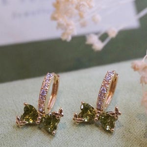 May include: A pair of gold hoop earrings with a green bow design. The hoops are set with clear cubic zirconia stones and the bows are accented with green stones.