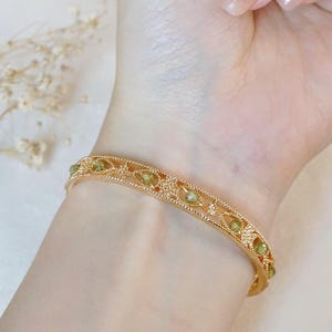 Pode incluir: Pulseira dourada com um delicado design de filigrana, adornada com pedras preciosas ovais verde claro. A pulseira é mostrada em um pulso, mostrando seus detalhes intrincados e seu estilo elegante. Uma joia.