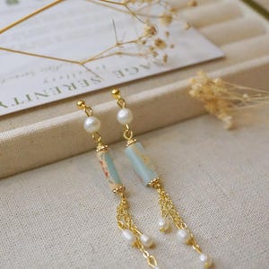 May include: A pair of dangle earrings with gold-colored chains and accents. Each earring features a light blue cylindrical bead with a floral design, a white pearl, and a chain of small white pearls.