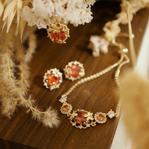 Puede incluir: Un conjunto de joyas doradas con collar, pendientes y colgante. El conjunto incluye piedras preciosas de color rojo anaranjado y cristales transparentes. Las joyas se exhiben sobre una superficie de madera oscura con flores secas en el fondo.