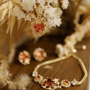 Op de afbeelding: Een goudkleurige sieradenset met een ovale, oranje-rode edelsteen hangerketting, bijpassende oorknopjes en een armband. De sieraden zijn geaccentueerd met kleine, heldere edelstenen en bloemendetails. De set wordt gepresenteerd op een houten oppervlak met gedroogde bloemen op de achtergrond.