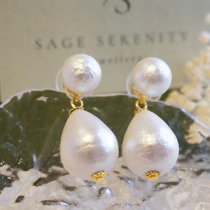 May include: A pair of white pearl drop earrings with gold-coloured accents. The earrings feature a small, round pearl at the top and a larger, teardrop-shaped pearl below. The earrings are displayed on a clear, textured surface.