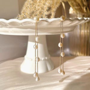 Könnte beinhalten: Elegante goldfarbene Ohrringe mit hängenden Perlen. Jeder Ohrring hat eine feine Kette, an der mehrere glänzende, cremefarbene Perlen befestigt sind. Die Ohrringe werden auf einer weißen, mehrstöckigen Tortenplatte präsentiert, um ihr anmutiges Design und ihren klassischen Stil hervorzuheben.