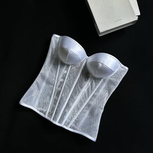 May include: White lace corset with satin cups and boning. The corset features a floral lace pattern and a structured silhouette. A book with the text "Better CLOTHING" is in the background.
