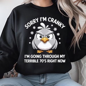 Op de afbeelding: Zwarte sweatshirt met de tekst "SORRY I'M CRANKY" en een cartoonvogel met een boze uitdrukking. De tekst "I'M GOING THROUGH MY TERRIBLE 70'S RIGHT NOW" staat eronder. Witte sneeuwvlokken omringen de vogel.