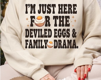 May include: A beige sweatshirt with the text "I'm just here for the deviled eggs & family drama." in black. The text is surrounded by two small stars and two deviled egg illustrations.