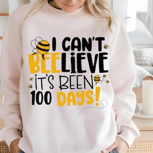 Op de afbeelding: Een lichtroze sweatshirt met de tekst "I Can't Bee-lieve It's Been 100 Days!" in zwart en geel. Het ontwerp bevat bijenillustraties. De sweatshirt heeft een ronde hals.