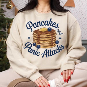 Peut inclure: Un sweat-shirt beige avec un graphique d'une pile de crêpes avec des myrtilles et une noix de beurre. Le texte "Pancakes and Panic Attacks" est imprimé en bleu sur le sweat-shirt.