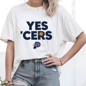 May include: White t-shirt with the text "YES 'CERS" in navy blue, featuring a basketball and the Pacers logo. The shirt has short sleeves and a relaxed fit, suitable for casual wear.