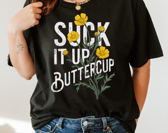 May include: Black t-shirt with the text "SUCK IT UP BUTTERCUP" in white, featuring yellow buttercup flowers and green leaves. The shirt is a casual style, suitable for everyday wear.