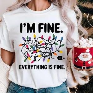 Puede incluir: Camiseta blanca con un gráfico negro de luces de Navidad enredadas y el texto "Estoy bien. Todo está bien."