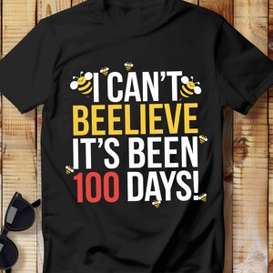 Op de afbeelding: Zwart T-shirt met een witte en gele afbeelding met de tekst "I can't beelieve it's been 100 days!" met bijenillustraties.