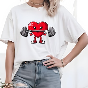 May include: White t-shirt featuring a red heart graphic lifting a barbell. The heart has a smiling face with eyelashes and red cheeks.