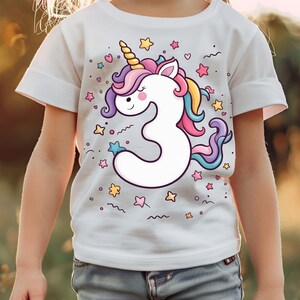 May include: White t-shirt featuring a colorful unicorn design with a number 3. The unicorn has a yellow horn, pink and purple mane, and a blue and yellow tail. The shirt is decorated with stars and hearts.