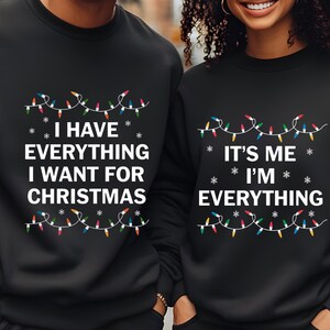 以下が含まれることがあります： クリスマスデザインの黒いスウェットシャツ。片方は「I HAVE EVERYTHING I WANT FOR CHRISTMAS」、もう片方は「IT'S ME I'M EVERYTHING」と書かれています。どちらもカラフルなライトと雪の結晶のデザインです。