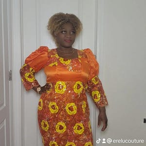May include: A woman wearing a long orange and yellow dress with a sequined floral pattern and fringe trim. The dress has a square neckline and puffed sleeves. The woman is standing in front of a white door.