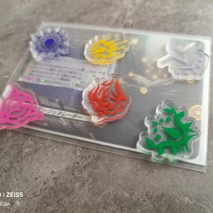 May include: Six clear acrylic tokens with different coloured symbols. The symbols are a blue sun, a yellow hand, a white lightning bolt, a red flame, a pink swirl, and a green leaf.