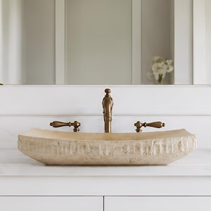 May include: A bathroom vanity with a beige stone vessel sink and bronze fixtures. The sink sits atop a white countertop and white cabinets with bronze hardware. A mirror is centered above the sink, and a white vase with a green flower is on the left.