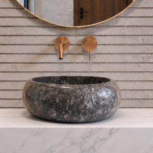 May include: A round, dark gray marble vessel sink sits on a white marble countertop. To the left is a black marble tumbler, and to the right is a small soap dish with a bar of soap. Above the sink are copper wall-mounted faucet fixtures.