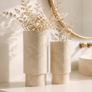 Könnte beinhalten: Zwei zylindrische Vasen aus beigem Travertin mit einer glatten, marmorierten Textur. Jede Vase verfügt über ein gestuftes Sockeldesign und enthält eine Ansammlung getrockneter, federartiger botanischer Stängel in hellen Beige- und Cremetönen.
