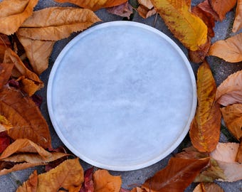 May include: A round, light blue serving tray with a textured surface. The tray is surrounded by fallen autumn leaves in shades of brown, orange, and yellow, creating a seasonal display. The tray is likely made of stone or ceramic.