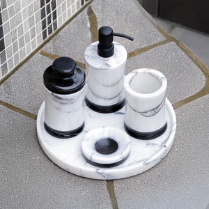 May include: A marble bathroom set includes a soap dispenser, tumbler, jar, and small dish, all with black accents. The set sits on a round tray, offering a cohesive and stylish design for bathroom organization.