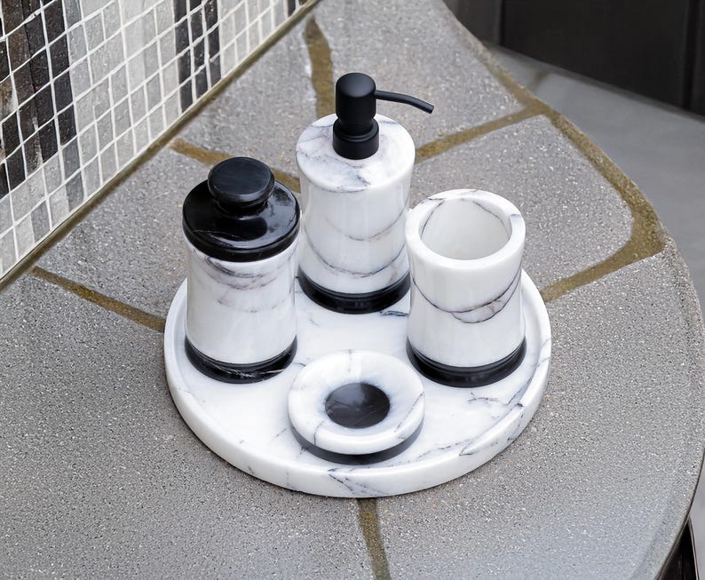 May include: A marble bathroom set includes a soap dispenser, tumbler, jar, and small dish, all with black accents. The set sits on a round tray, offering a cohesive and stylish design for bathroom organization.