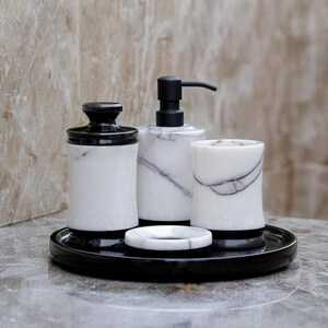 May include: A bathroom set featuring a black and white marble design. The set includes a soap dispenser, a jar with a lid, a tumbler, and a soap dish, all resting on a black tray. The soap dispenser has a black pump.