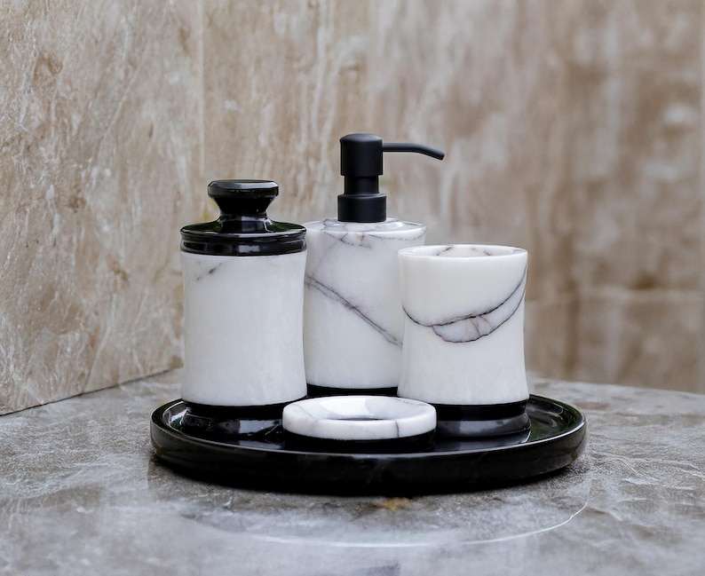May include: A bathroom set featuring a black and white marble design. The set includes a soap dispenser, a jar with a lid, a tumbler, and a soap dish, all resting on a black tray. The soap dispenser has a black pump.